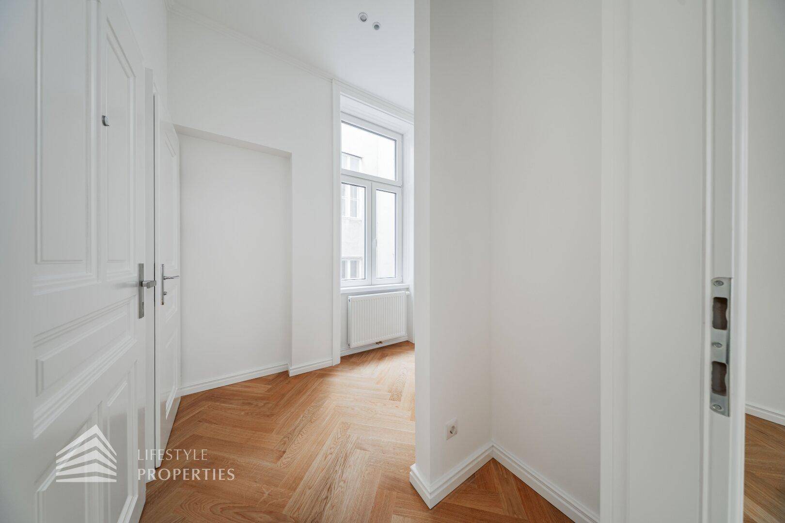 Schmaler Flur mit Fischgrätparkett und einem Fenster, das Tageslicht hereinlässt.