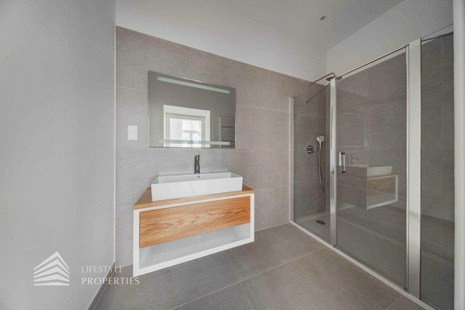 Modernes Badezimmer mit großformatigen grauen Fliesen, elegantem Waschtisch und Glasdusche.