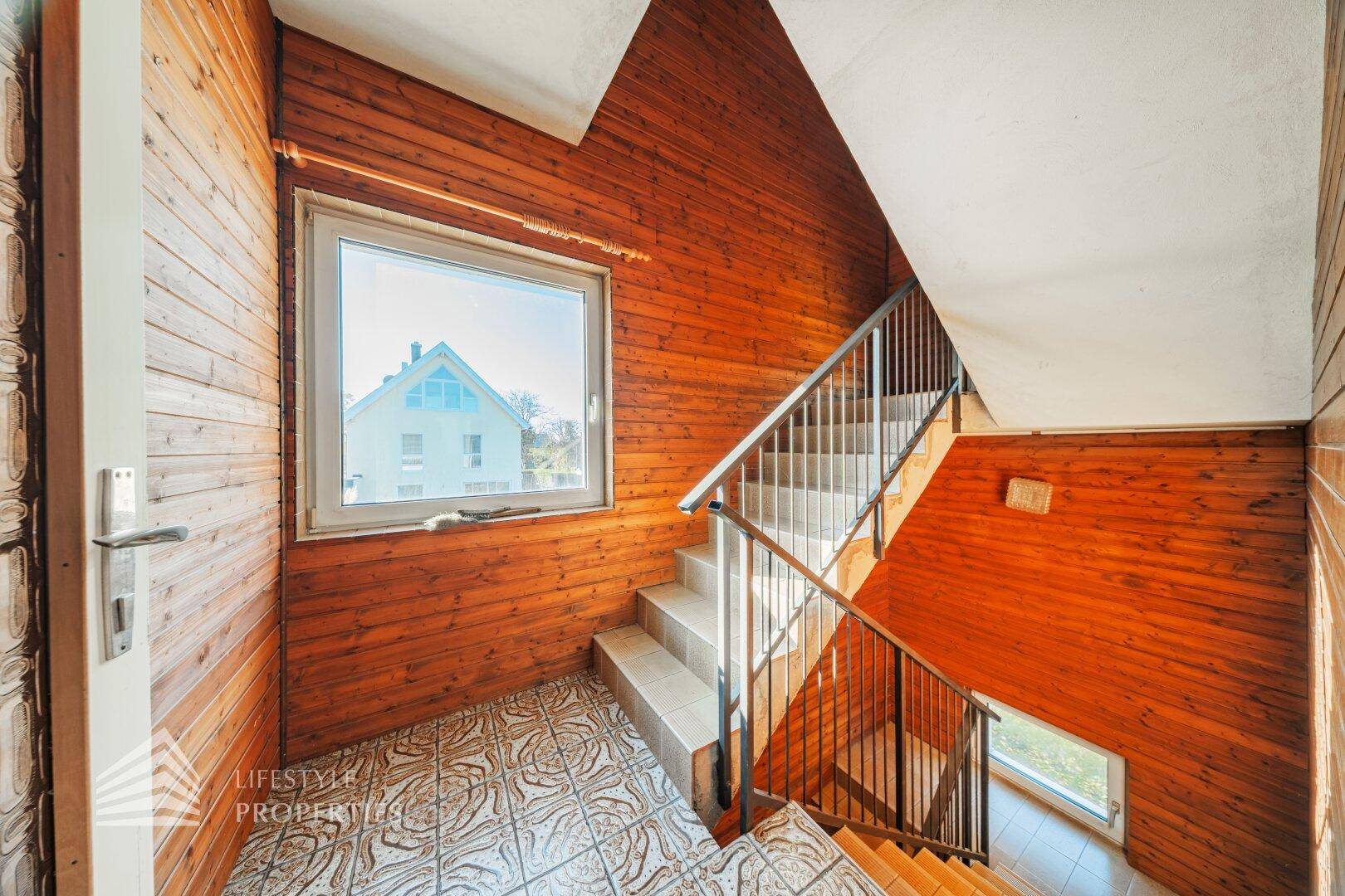 Treppenhaus mit Fenster, Metallgeländer und Holzverkleidung, bietet viel Tageslicht.