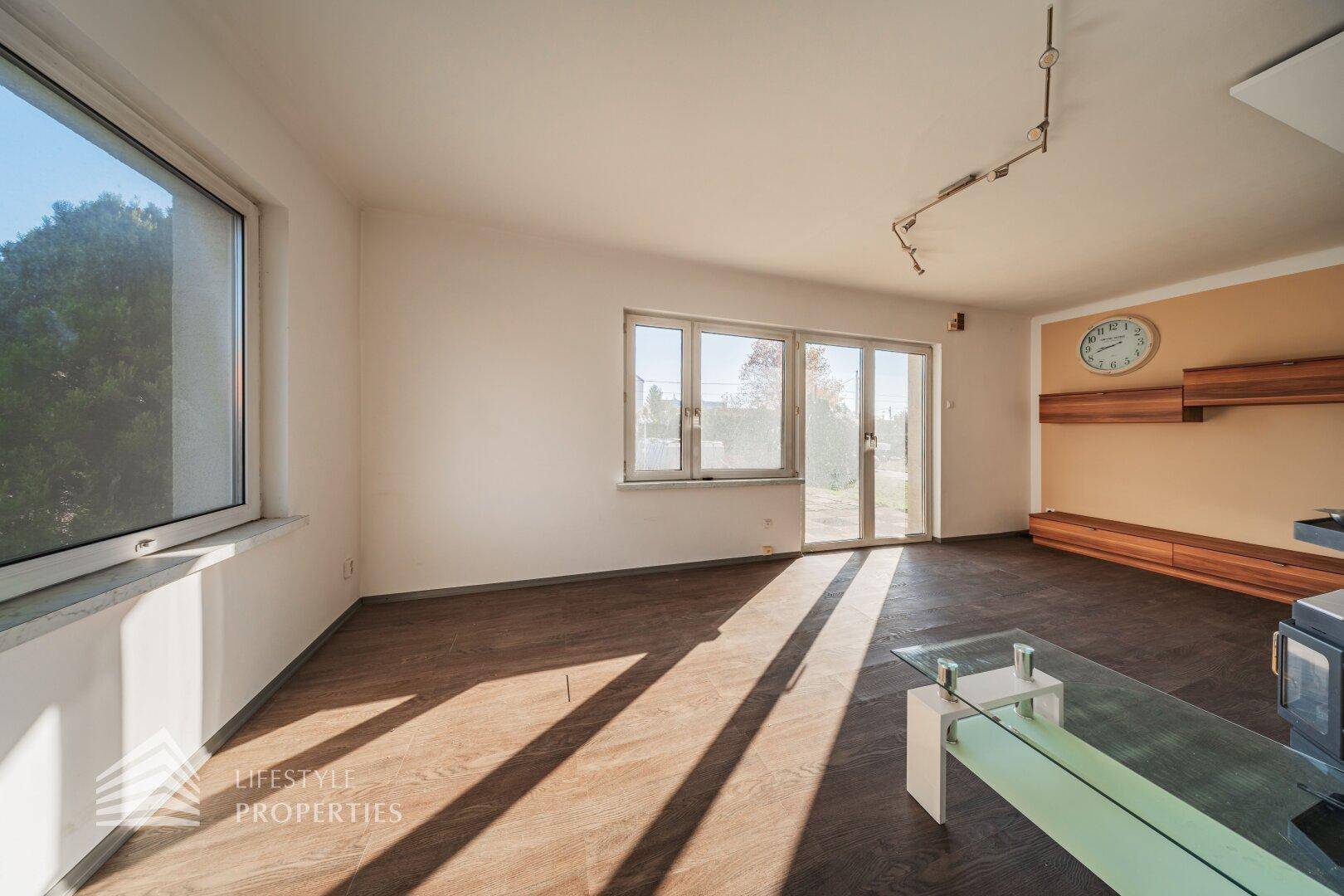 Geräumiges Wohnzimmer mit Holzboden, großen Fenstern und Wandregalen.