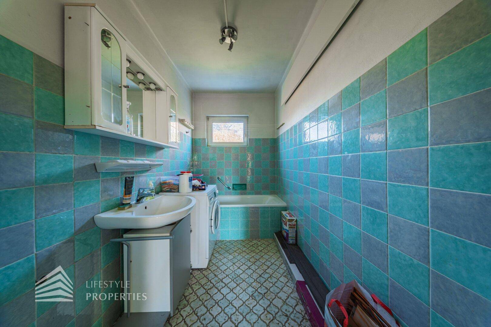 Badezimmer mit blauen und grauen Fliesen, Waschbecken, Badewanne und Waschmaschine.