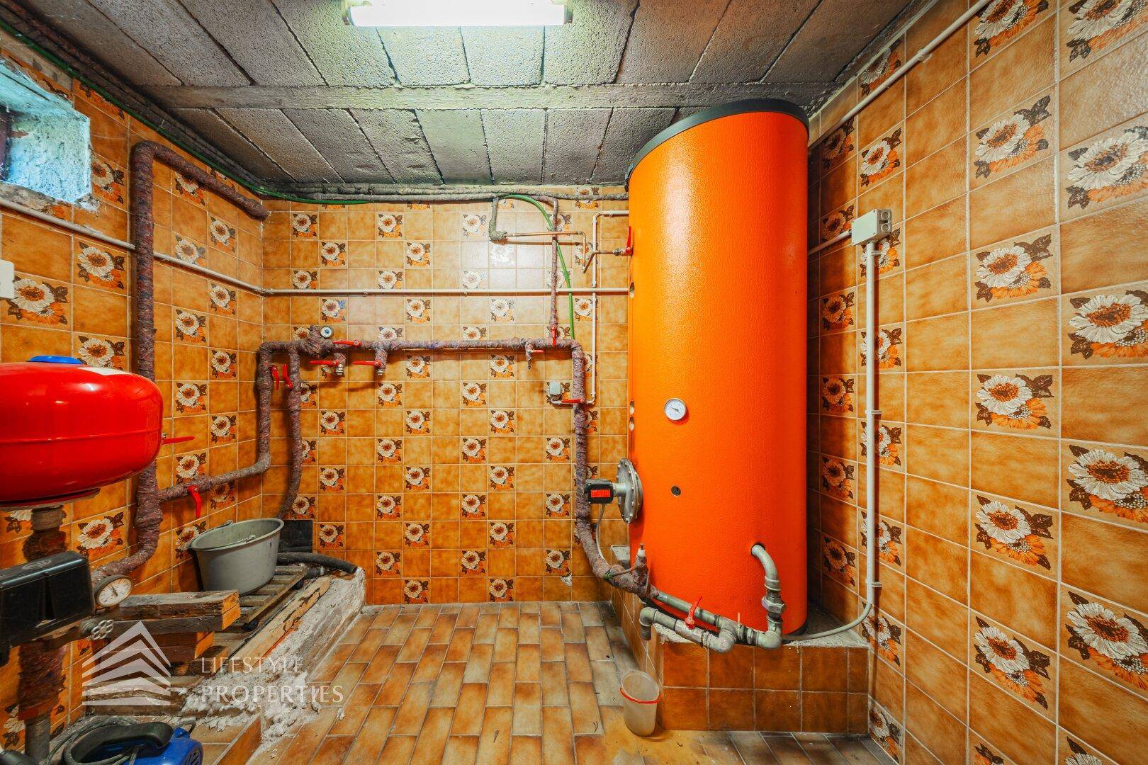 Heizungsraum mit großem Warmwasserspeicher, Rohren und gefliesten Wänden.