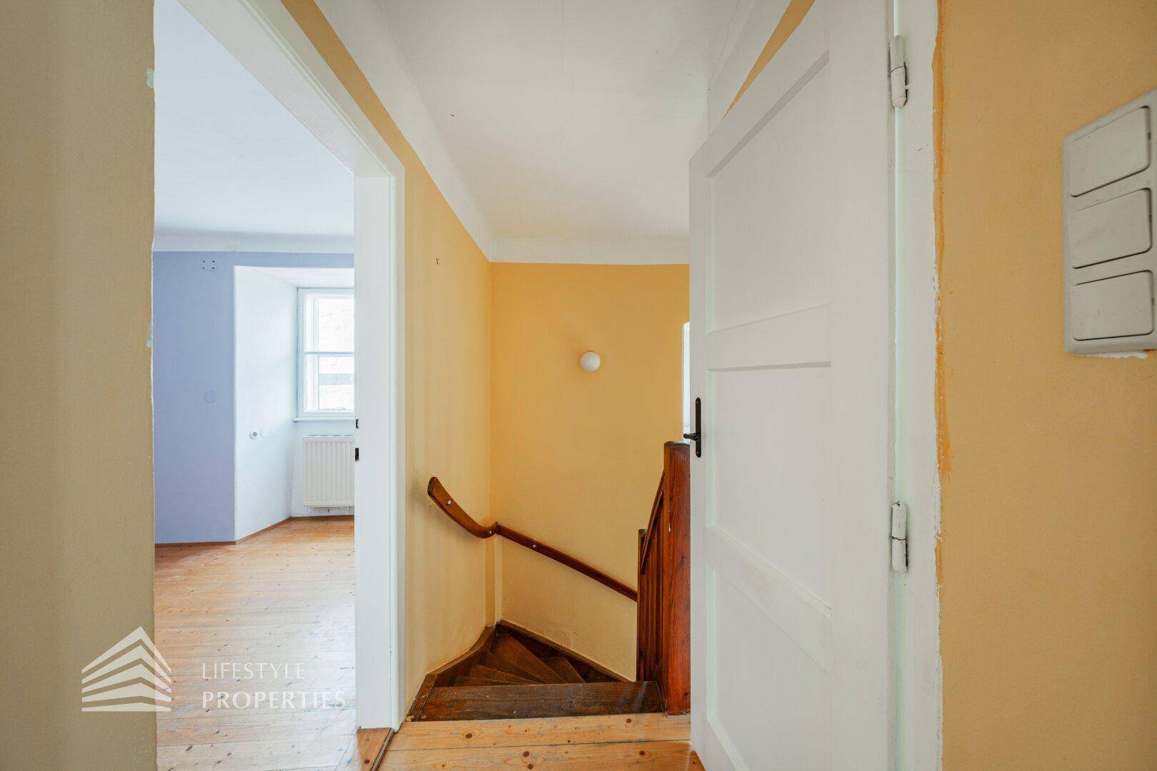 Flur mit Holztreppe und Zugang zu einem hellen Zimmer mit Holzboden.