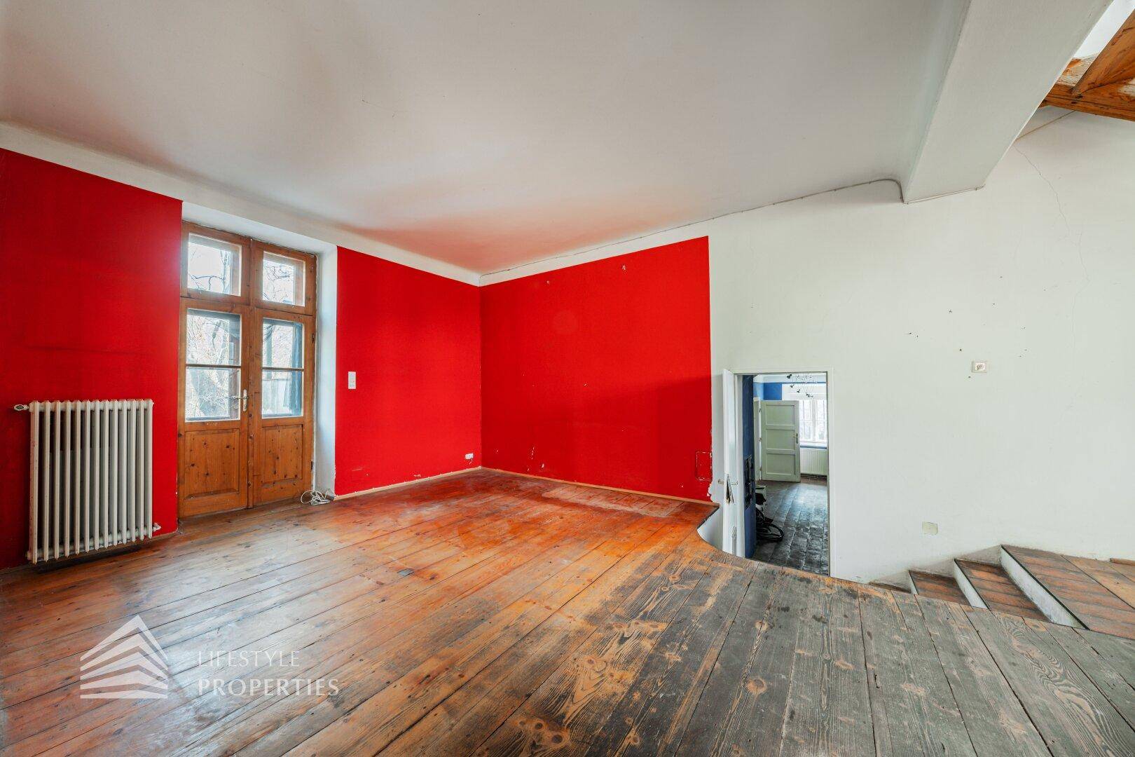 Geräumiges Zimmer mit Holzdielenboden, roten Wänden und Zugang nach draußen.