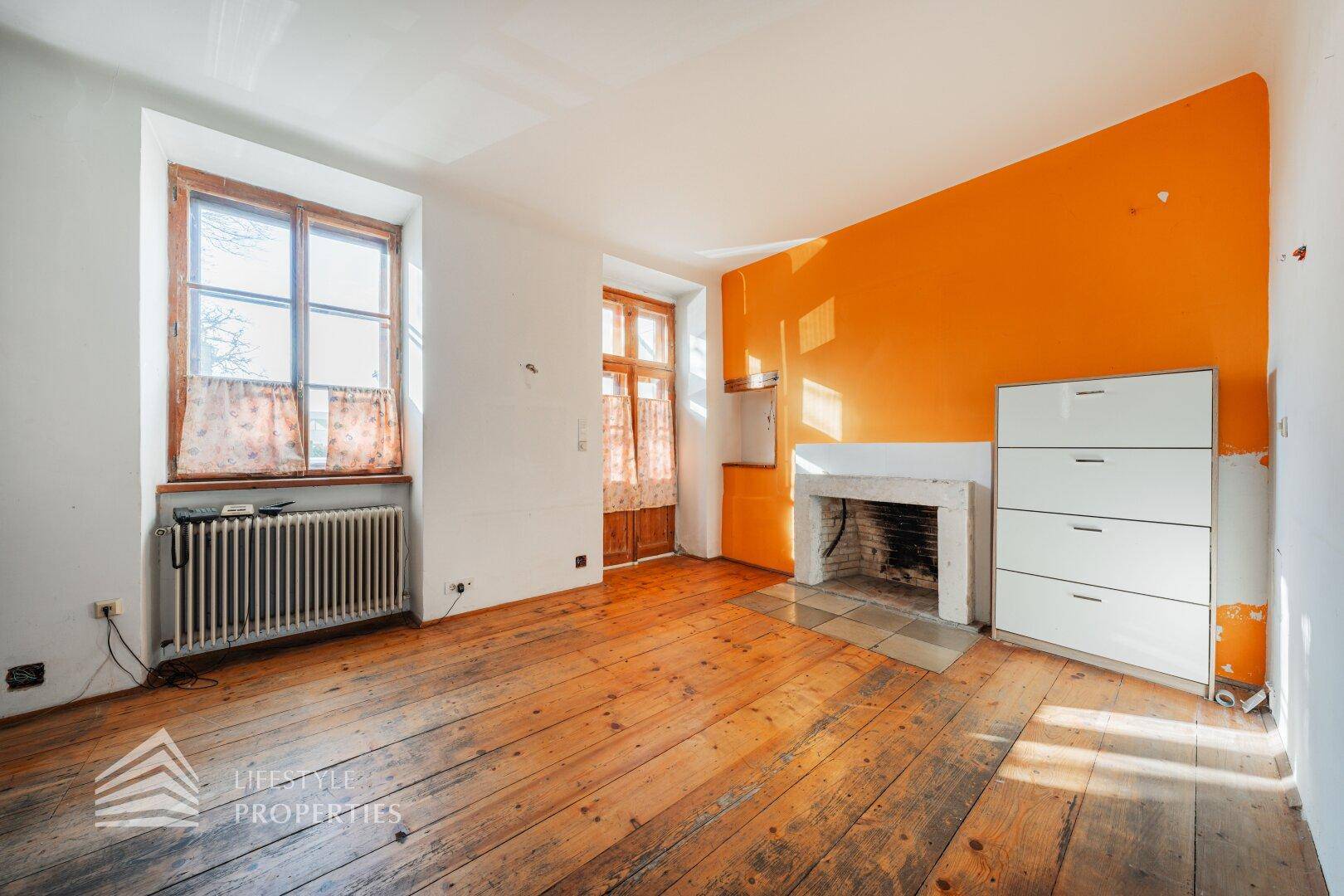 Heller Wohnbereich mit Holzdielenboden, Kamin und orangefarbener Akzentwand.