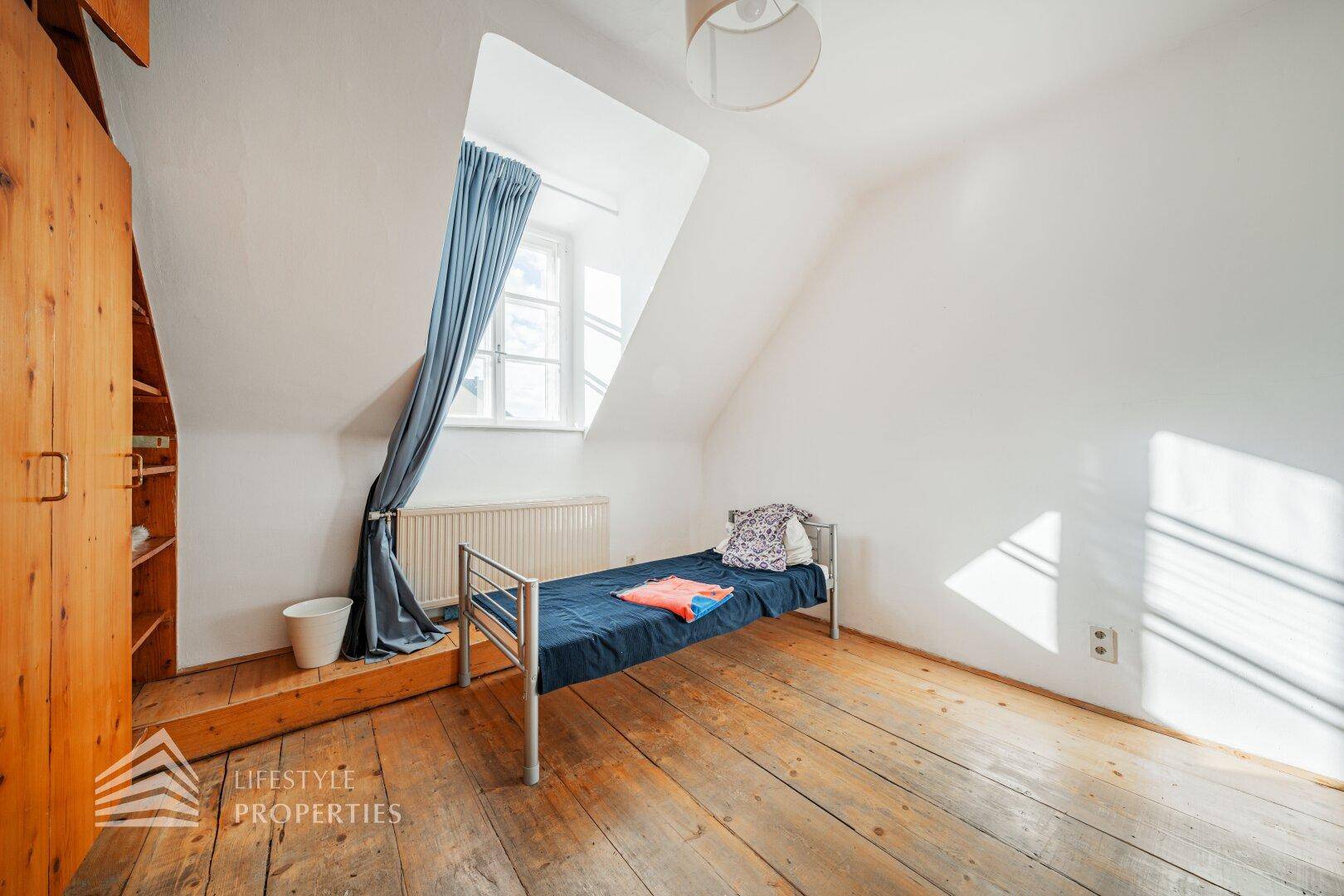 Helles Zimmer im Dachgeschoss mit Holzdielenboden, Dachschrägen und Fenster.