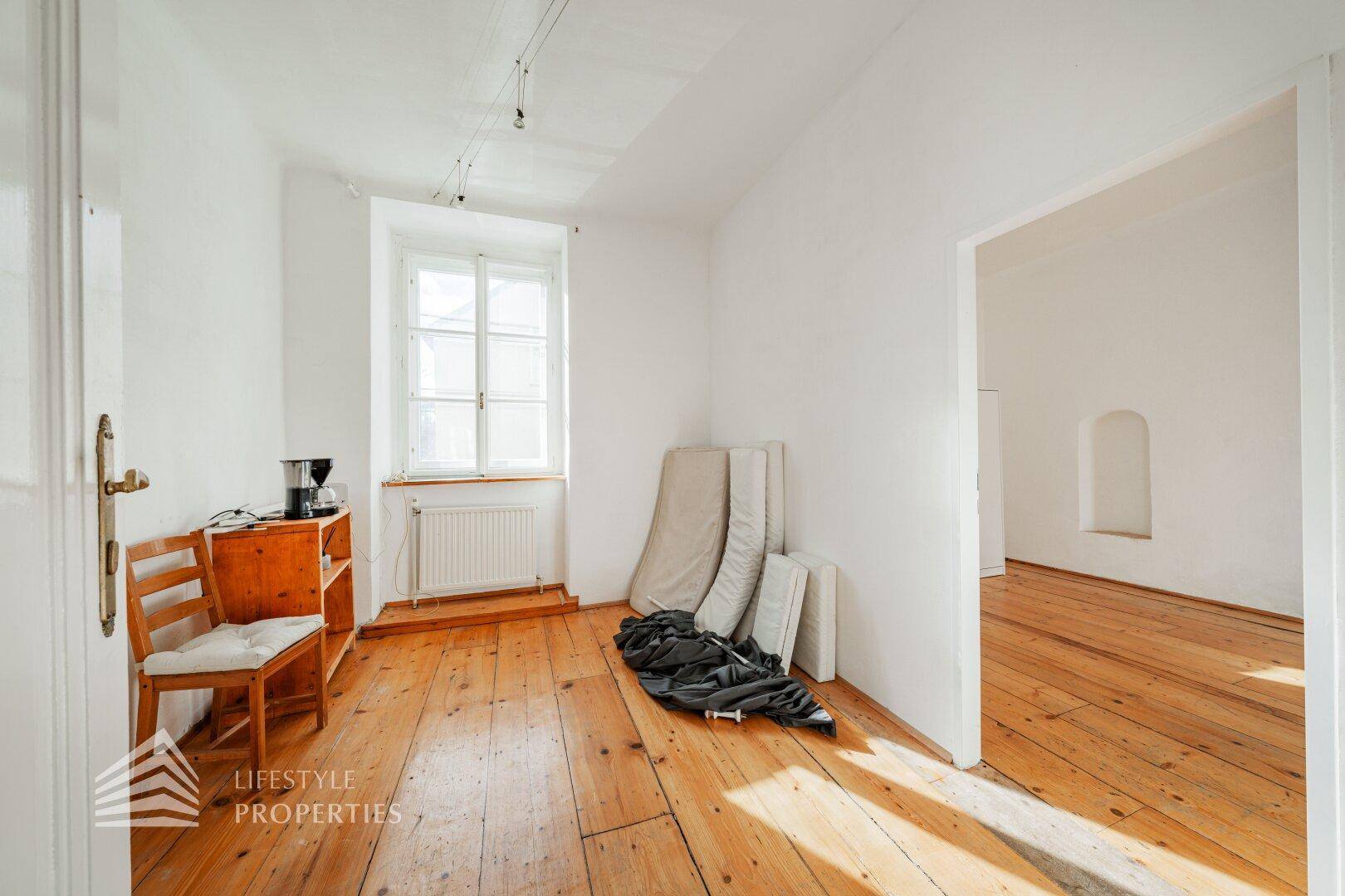 Helles Zimmer mit Holzdielenboden, weiß gestrichenen Wänden und großem Fenster.