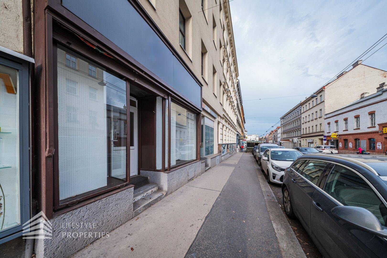 Seitenansicht des Gebäudes entlang einer städtischen Straße mit parkenden Autos und weiteren Gebäuden im Hintergrund.