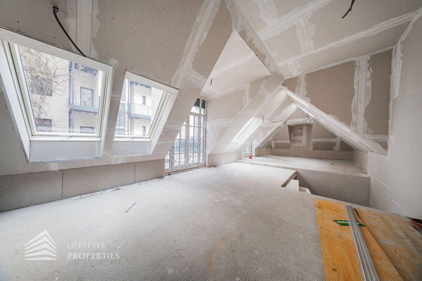 Dachgeschossraum im Rohbau mit schrägen Decken, Dachfenstern und Zugang zu einem Außenbereich.
