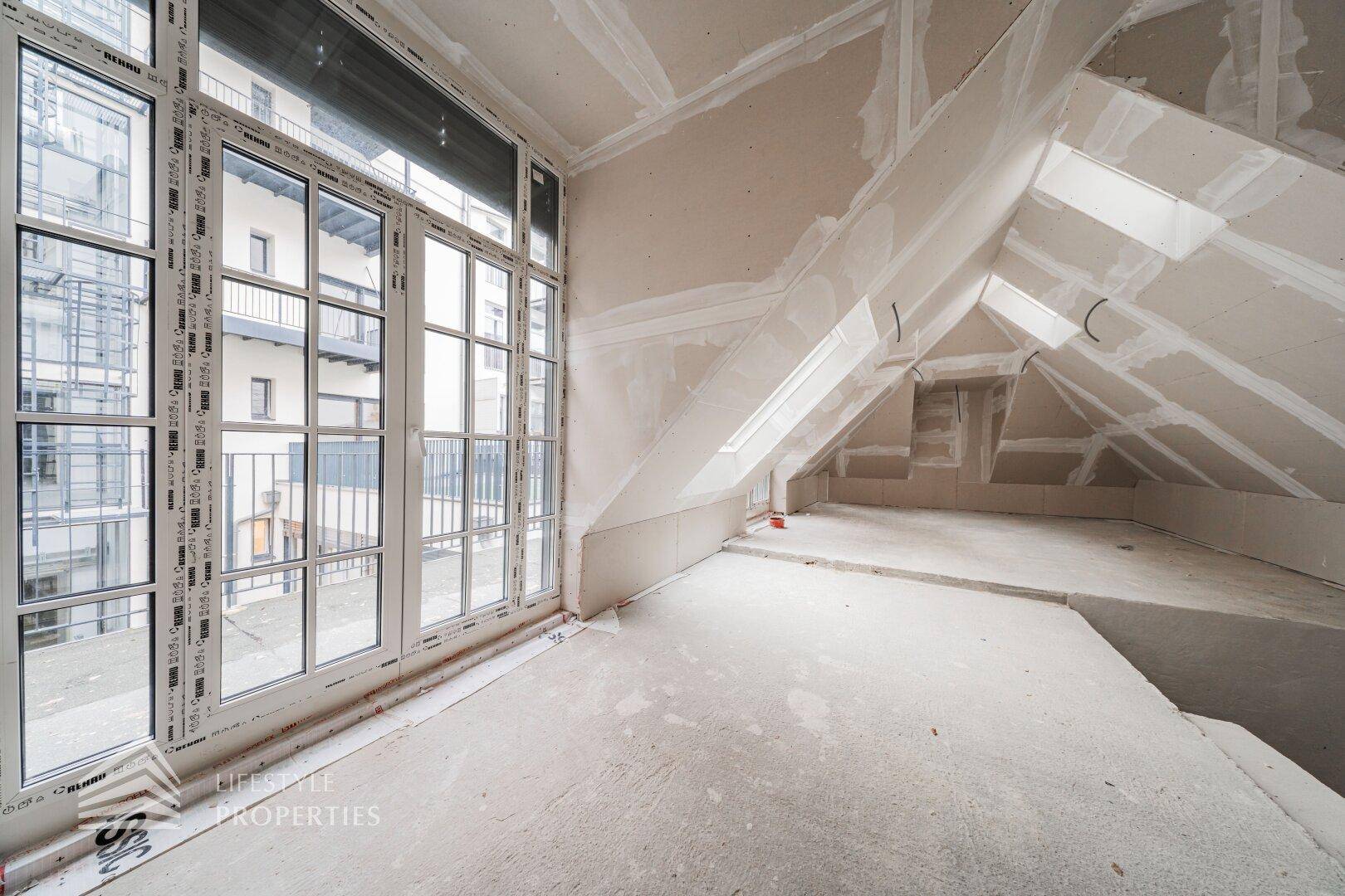 Dachgeschossraum im Rohbau mit schrägen Decken und großen Fenstern, die viel Licht hereinlassen.