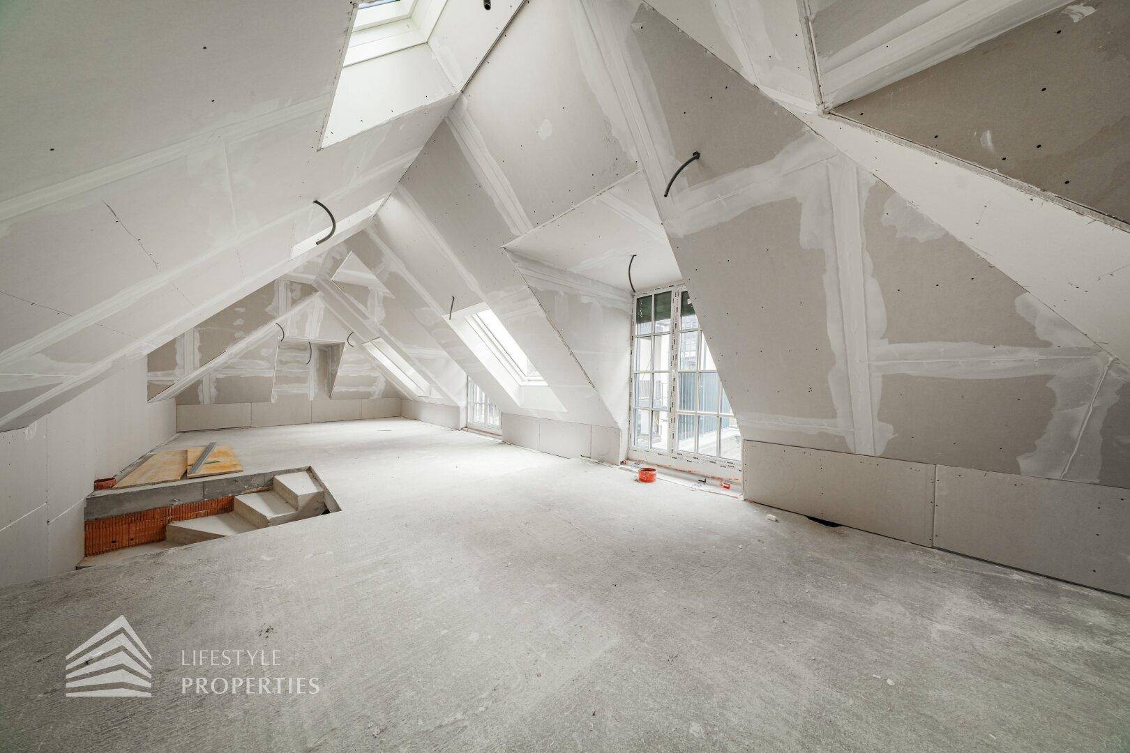 Geräumiger Dachgeschossraum im Rohbau mit mehreren Dachfenstern und schrägen Wänden.