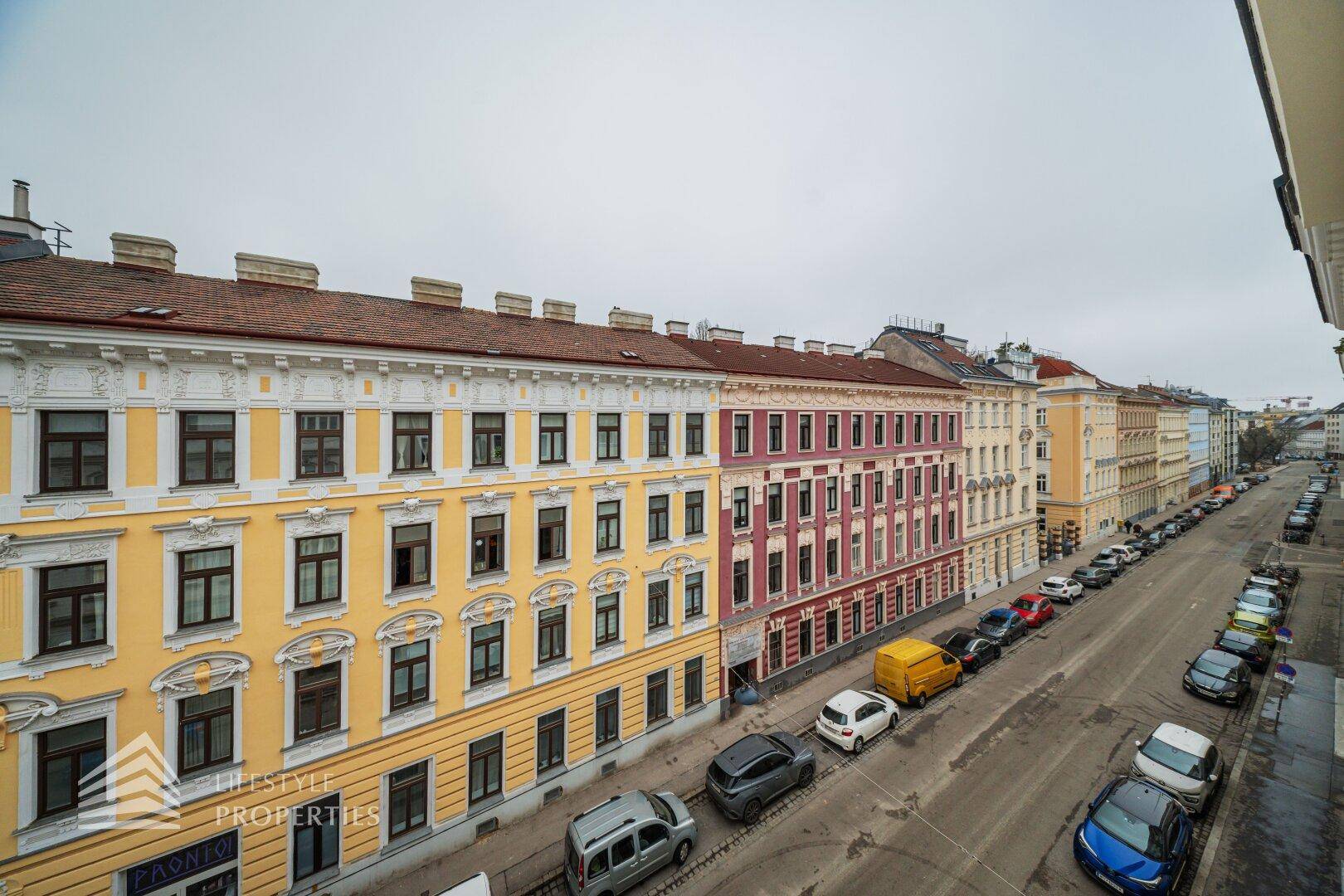 Weitläufiger Blick über eine städtische Straße mit farbenfrohen Altbauten und parkenden Fahrzeugen.