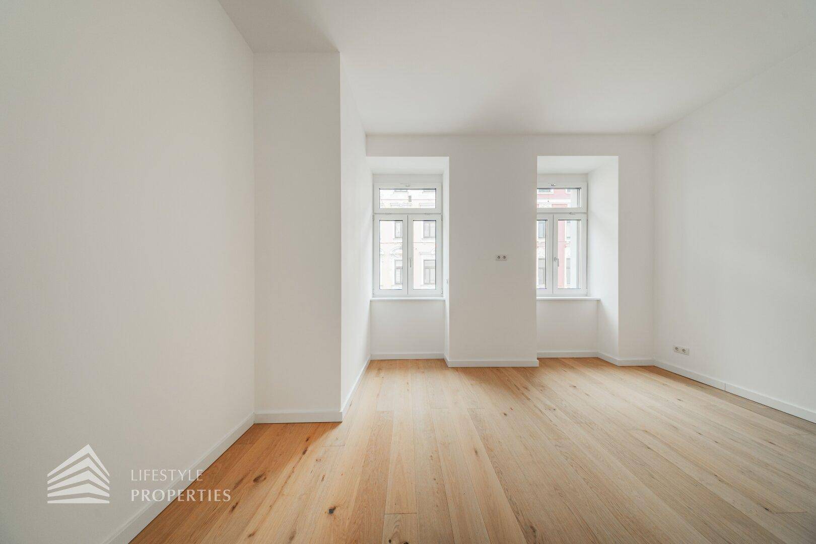 Leeres Zimmer mit hellem Parkettboden und zwei klassischen Fenstern, ideal als Schlaf- oder Arbeitszimmer.