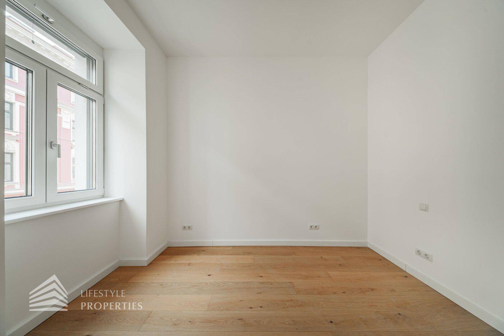 Helles, unmöbliertes Zimmer mit Holzboden und einem Fenster, das viel Tageslicht hereinlässt.