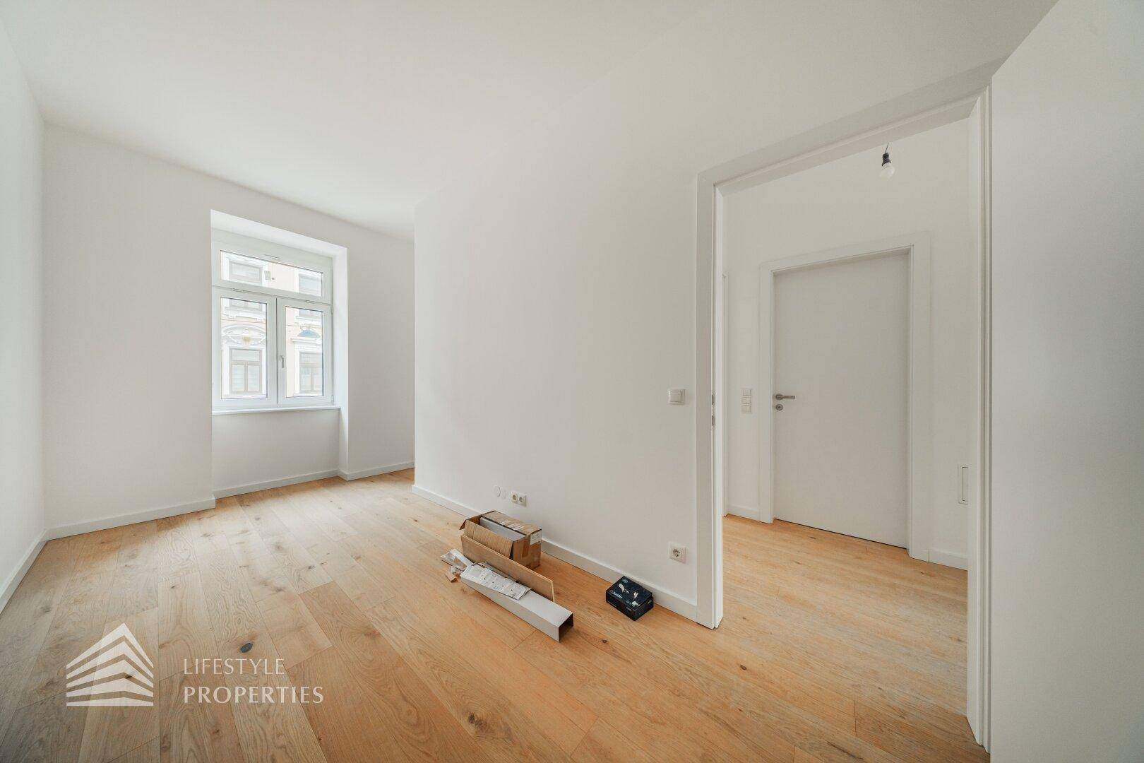 Unmöbliertes Zimmer mit hellem Parkettboden und einem Fenster, das den Raum mit Tageslicht füllt.