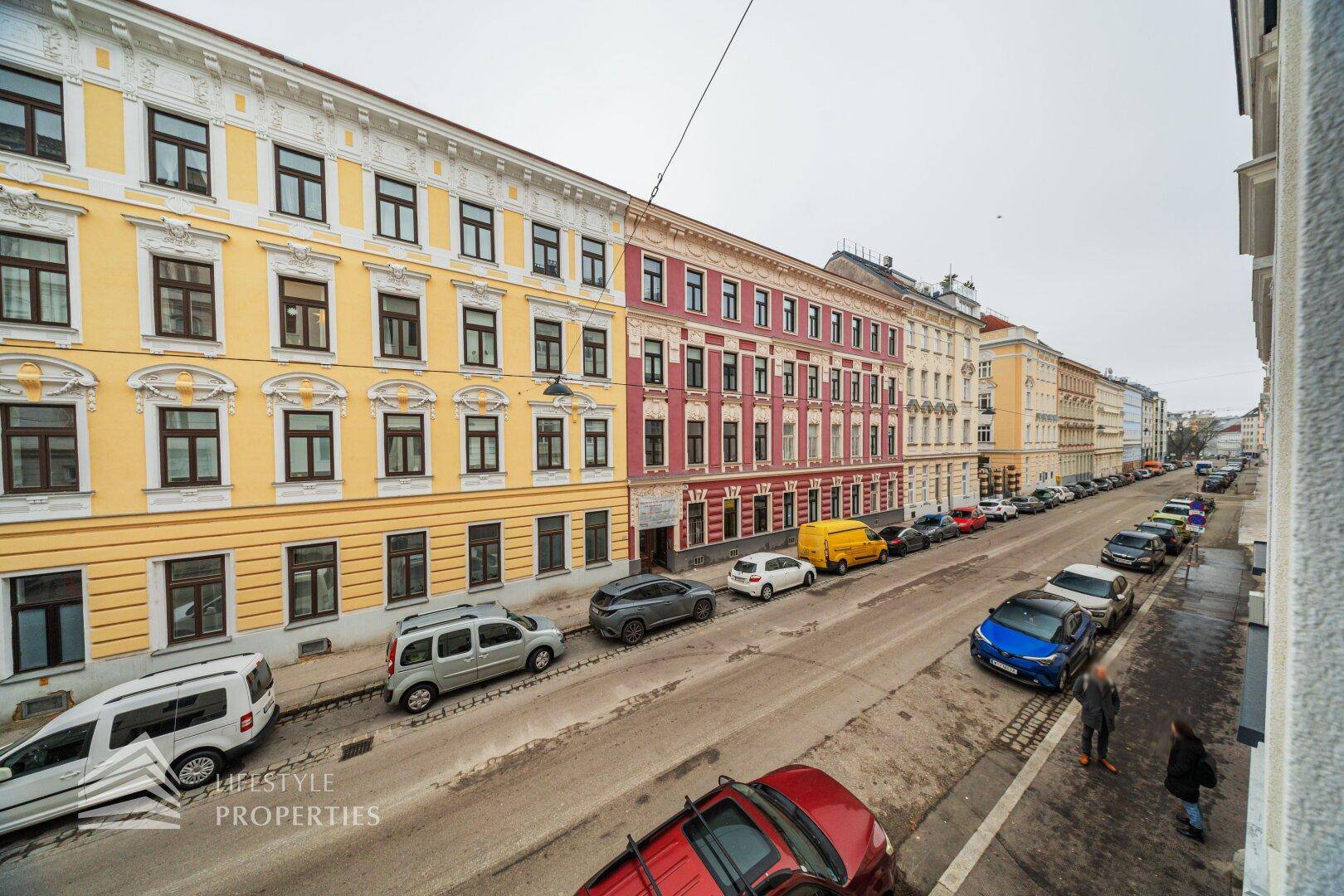 Blick von oben auf eine belebte Stadtstraße mit parkenden Autos und historischen Gebäudefassaden.