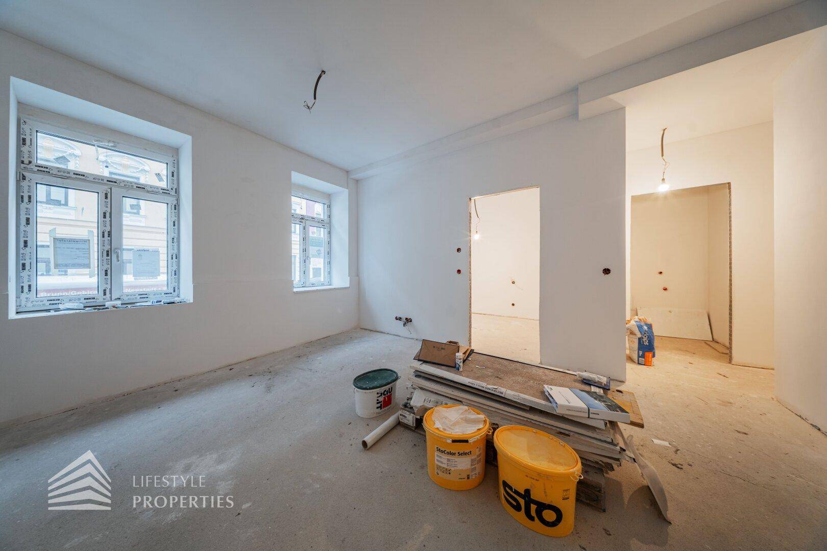 Heller Raum im Rohbauzustand mit zwei Fenstern und Baueimern auf dem Betonboden.