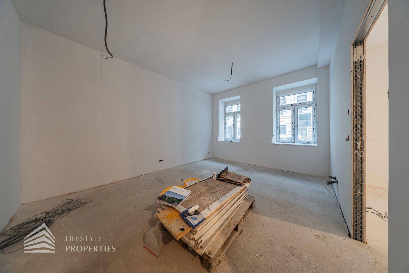 Leerer Raum im Rohbauzustand mit zwei Fenstern und Baumaterialien auf dem Betonboden.