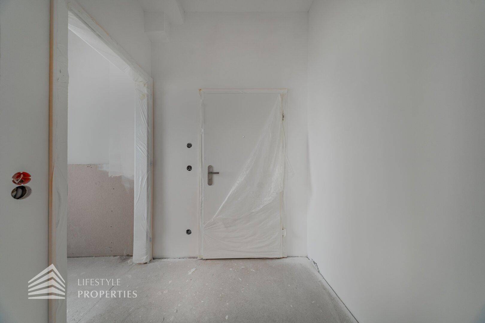 Eingangsbereich im Rohbau mit weißer, folierter Tür und unfertigen Wänden und Boden.