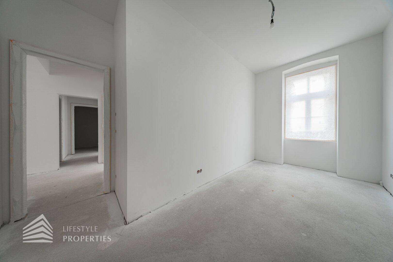 Unfertiger Raum mit Fenster und Zugang zu einem weiteren Zimmer im Rohbauzustand.
