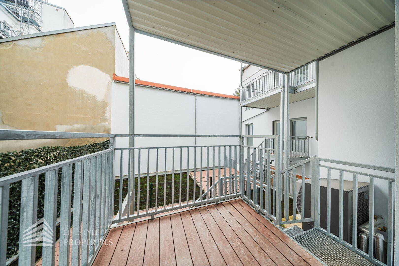 Balkon mit Holzboden und Metallgeländer, umgeben von Gebäudefassaden im Innenhof.