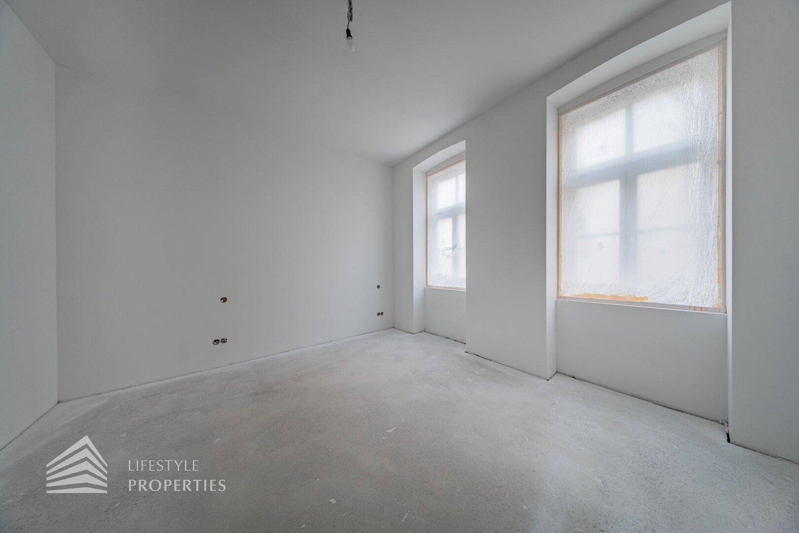 Leerer Raum im Bauzustand mit zwei Fenstern und Betonboden, bereit für den Innenausbau.