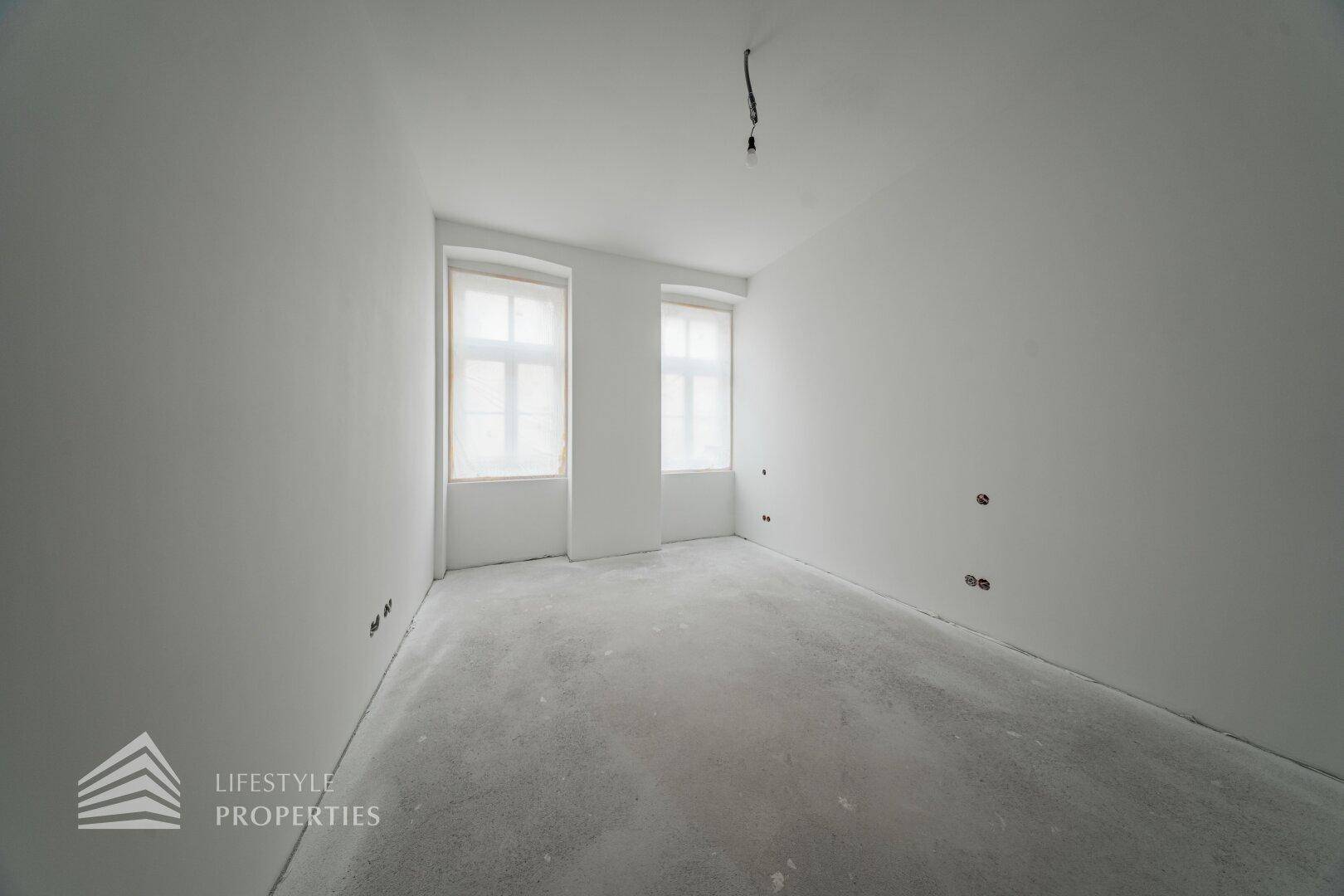 Leerer Raum im Bauzustand mit zwei Fenstern und Betonboden, bereit für den Innenausbau.
