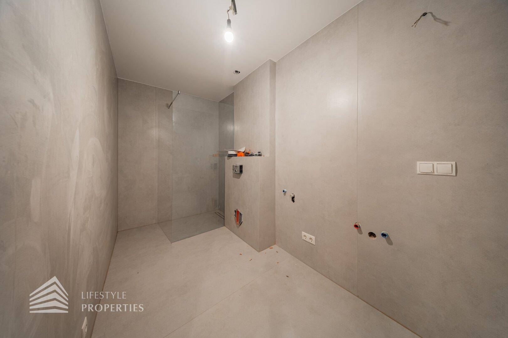 Unfertiges Badezimmer mit grauen Fliesen und vorbereiteten Anschlüssen für eine moderne Dusche.