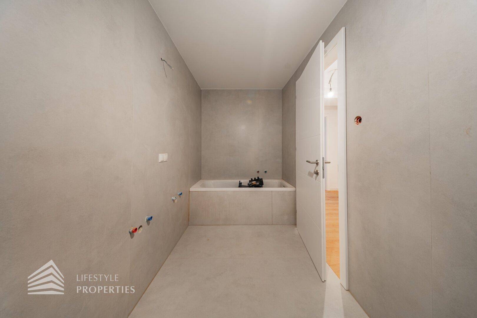 Unfertiges Badezimmer mit grauen Fliesen und vorbereiteten Anschlüssen für eine Badewanne.
