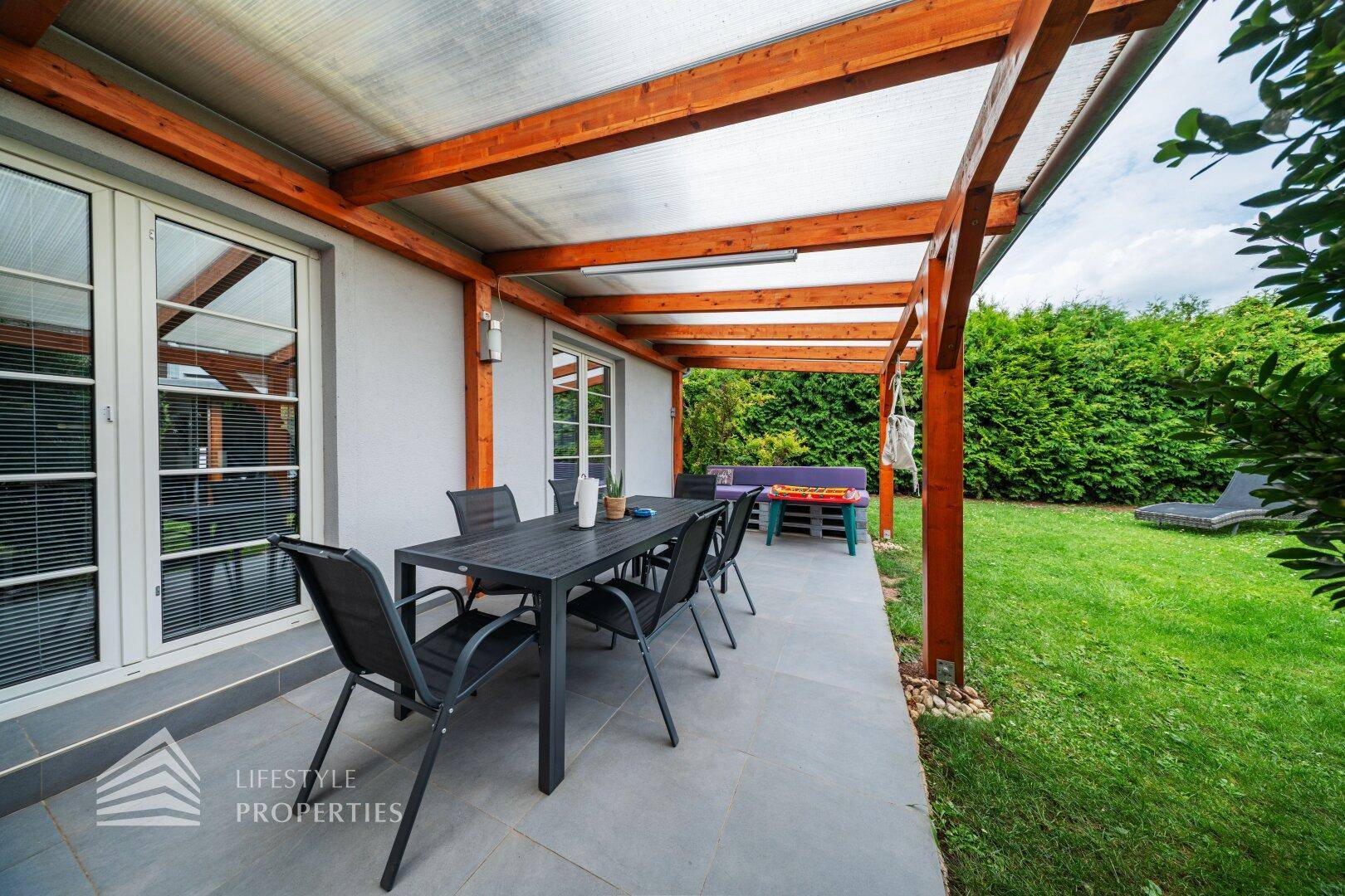Überdachte Terrasse mit Esstisch und Stühlen, umgeben von einem gepflegten Rasen und Hecken.