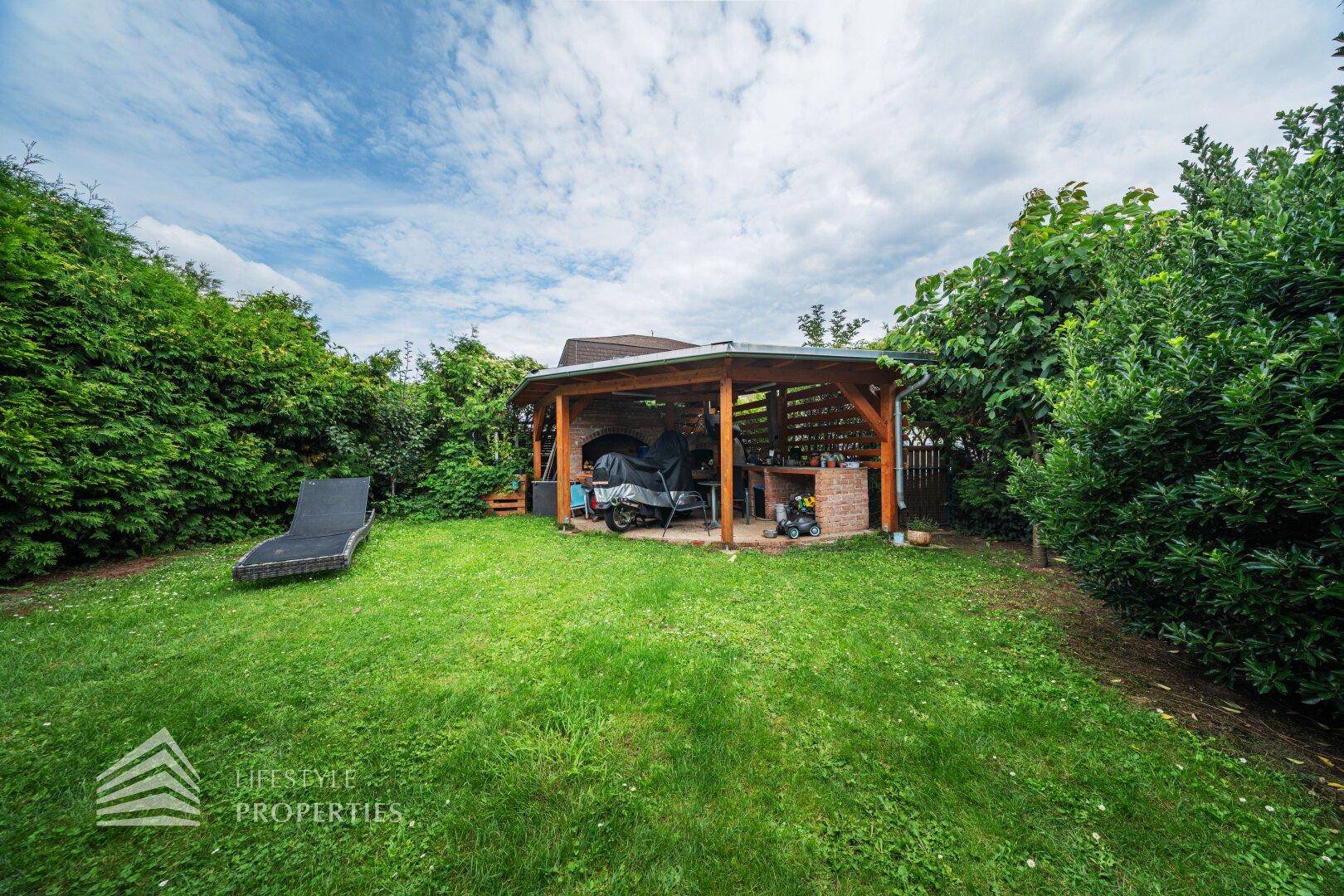 Großer Garten mit Rasenfläche, einer Holzpergola und einem Liegestuhl zum Entspannen.