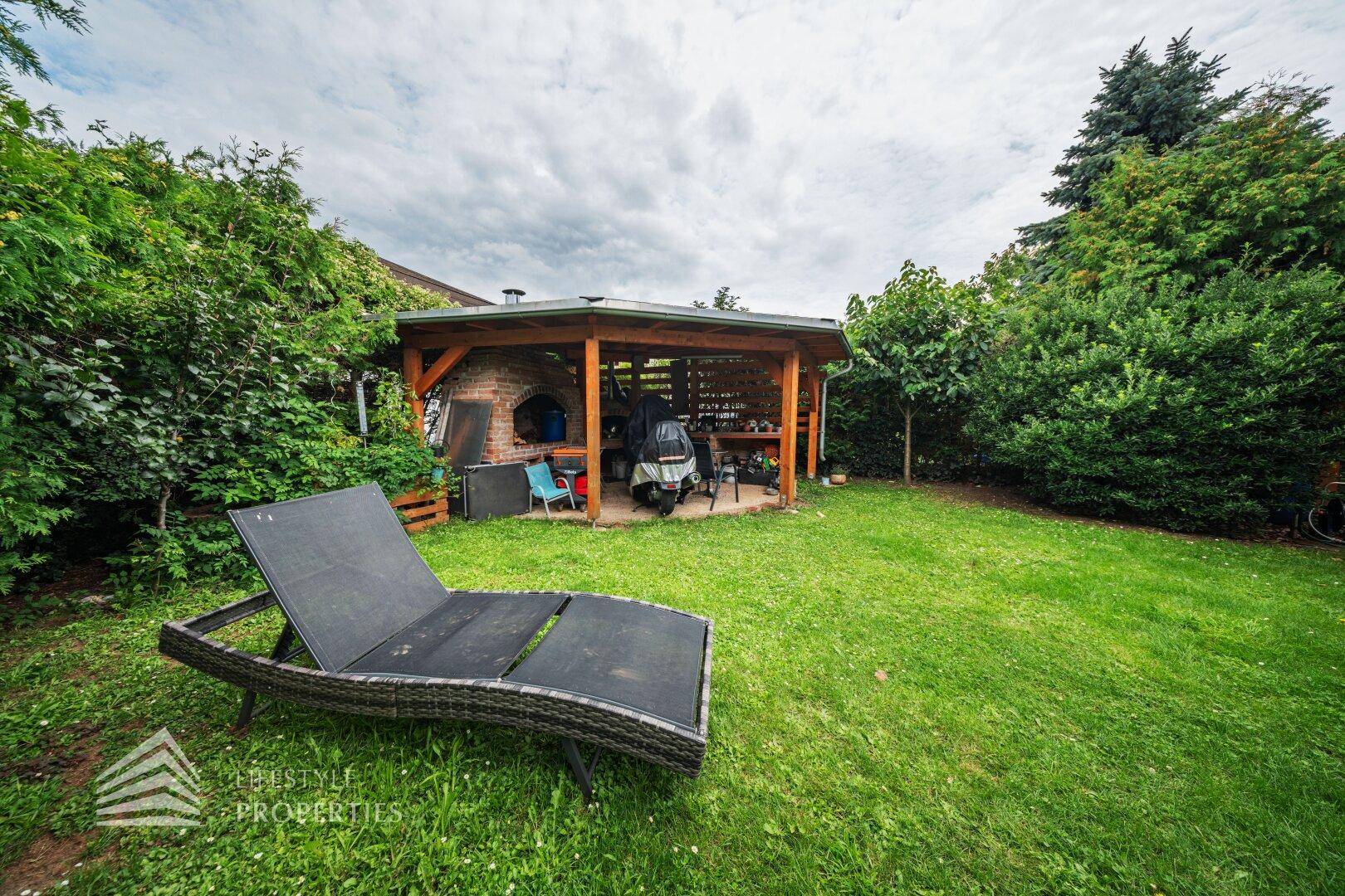 Gepflegter Garten mit Rasenfläche, einer Holzpergola mit Grillbereich und einem bequemen Liegestuhl.