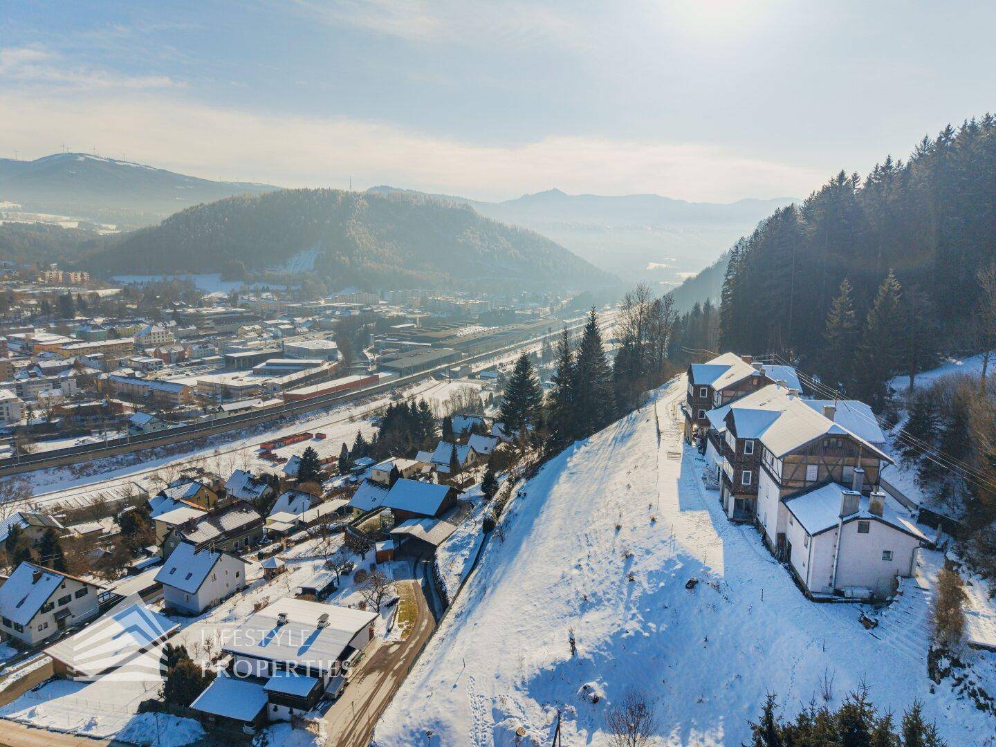 Panoramablick auf das Anwesen am Hang, umgeben von verschneiten Bergen und einem Tal mit Siedlung.
