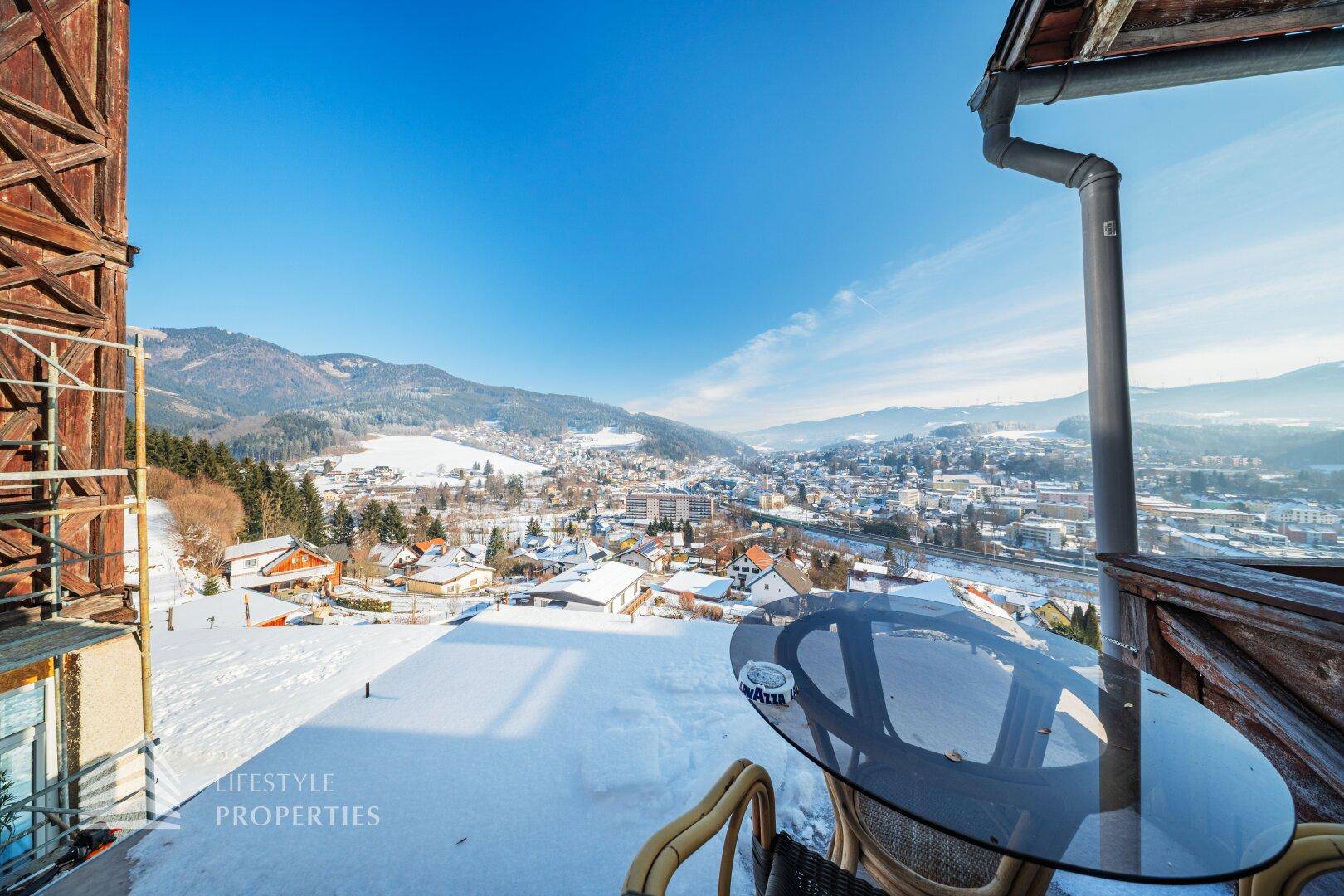 Balkon mit Sitzgelegenheiten und weitem Blick über eine verschneite Stadt und Berge.