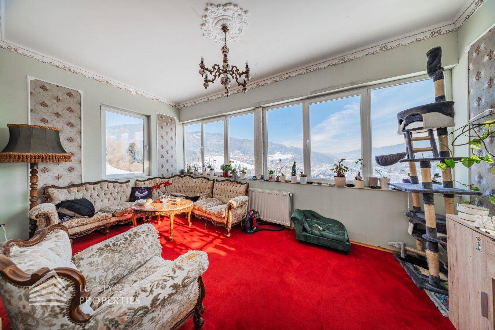 Wohnzimmer mit traditionellen Sofas, rotem Teppich und großen Fenstern mit Bergblick.
