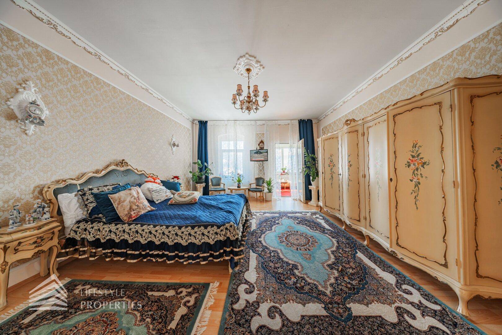 Schlafzimmer mit großem blauem Bett, bemalten Kleiderschränken und einem gemusterten Teppich auf Holzboden.