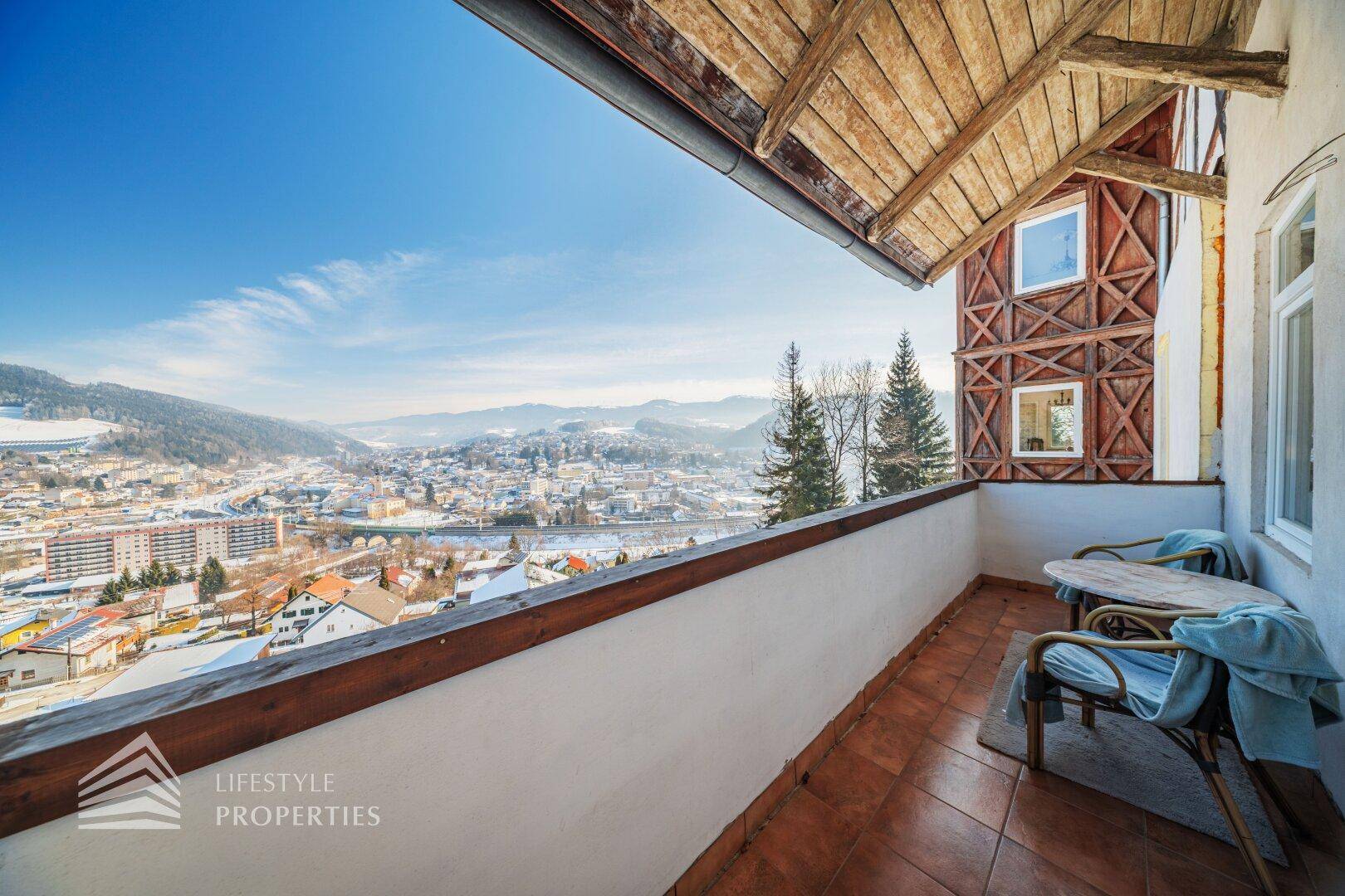 Balkon mit Fliesenboden, Holzgeländer und Panoramablick auf die verschneite Stadt und die Berge.