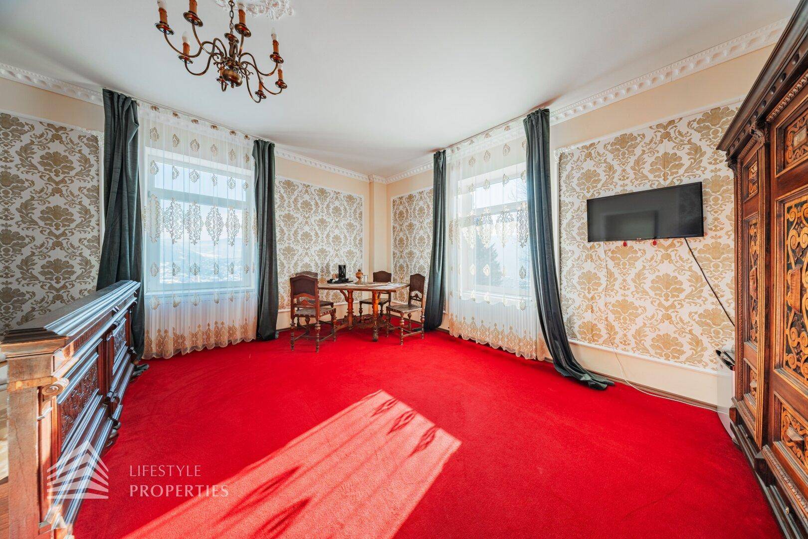 Großes Schlafzimmer mit rotem Teppich, traditionellen Möbeln und einem Wandfernseher, hell durch große Fenster.