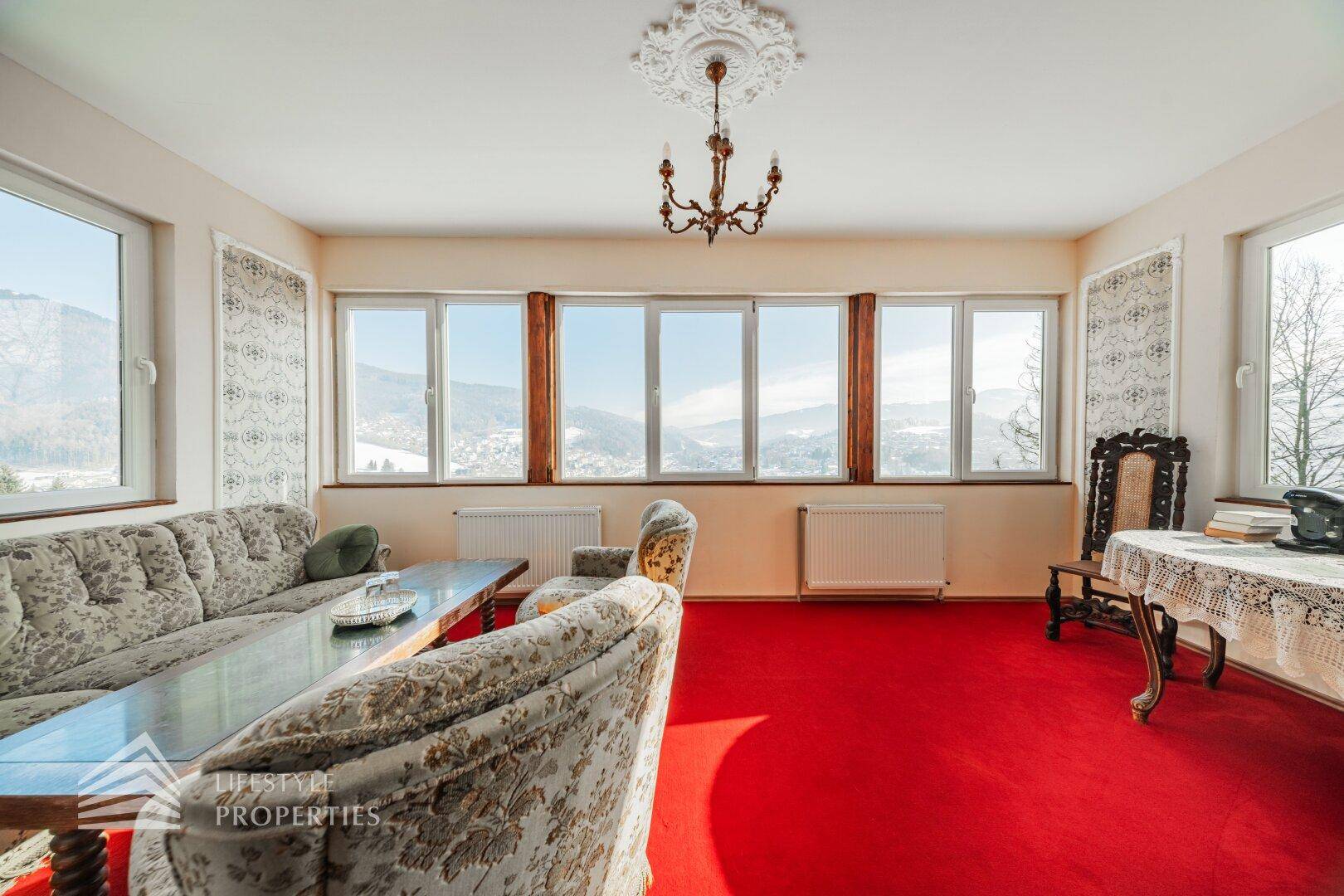 Gemütliches Wohnzimmer mit gemusterten Sofas, rotem Teppich und Panoramafenstern mit Bergblick.