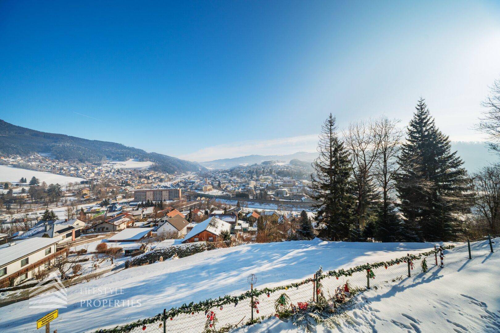 Weitläufiger Blick über eine verschneite Winterlandschaft mit einem Dorf im Tal unter strahlend blauem Himmel.