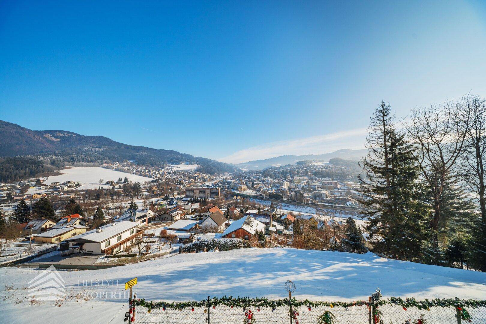 Panoramablick auf eine verschneite Berglandschaft und eine Siedlung im Tal an einem sonnigen Wintertag.