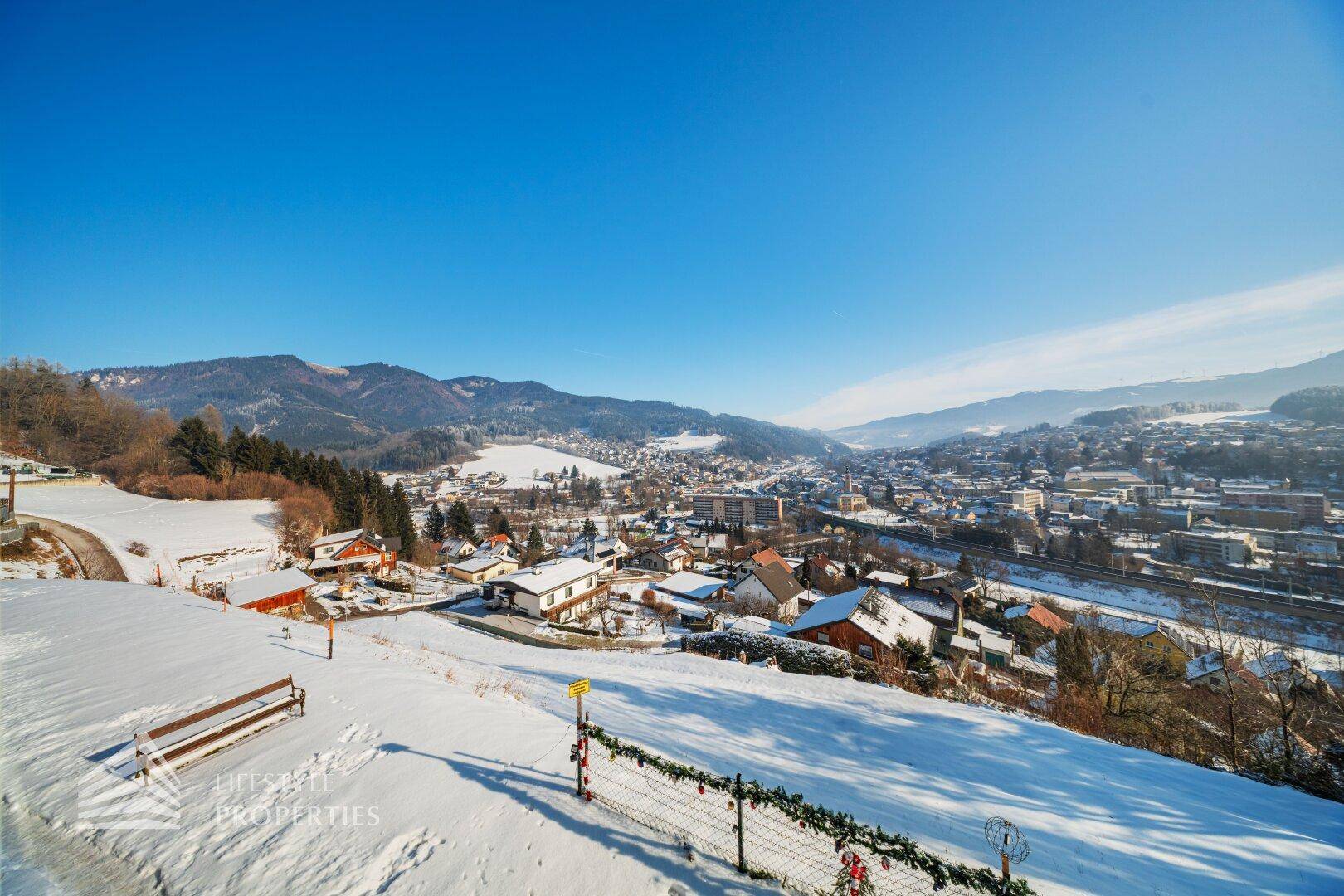 Malerischer Ausblick auf eine verschneite Landschaft mit einem Dorf und einer Bank im Vordergrund.