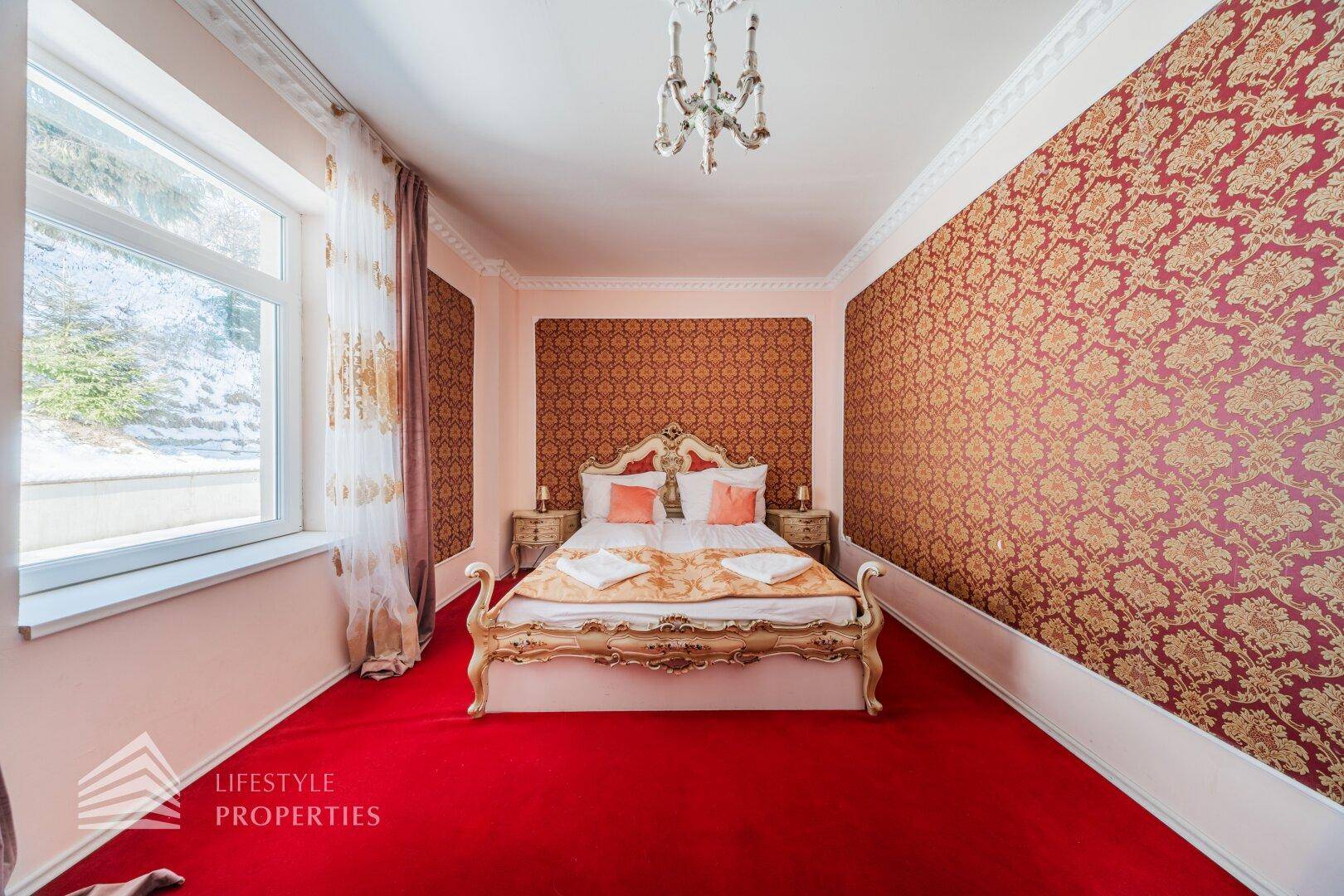 Elegantes Schlafzimmer mit Doppelbett, rotem Teppich und auffälliger rot-goldener Tapete.