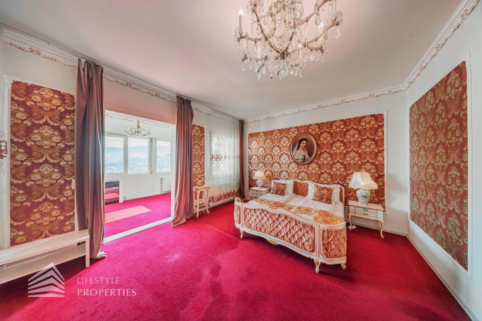Elegantes Schlafzimmer mit rotem Teppich, Kronleuchter und Zugang zu einem weiteren Raum mit Bergblick.