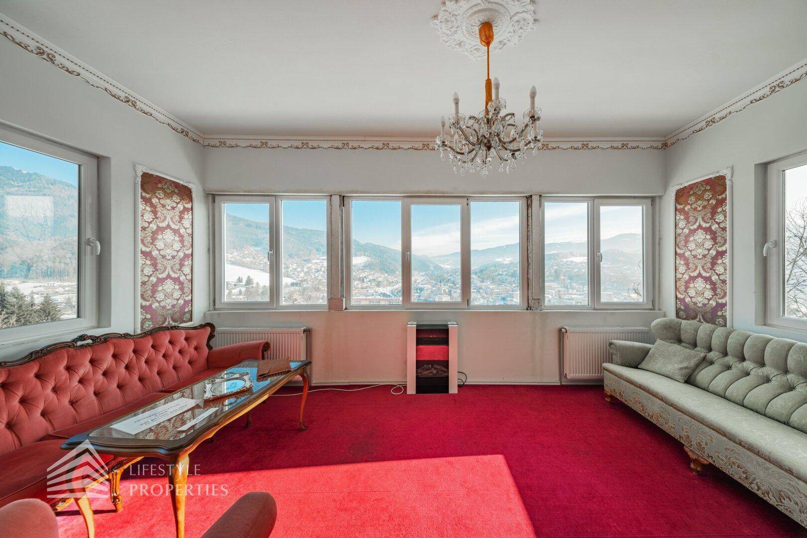 Wohnzimmer mit rotem Teppich, klassischen Sofas und großen Fenstern mit Bergblick.