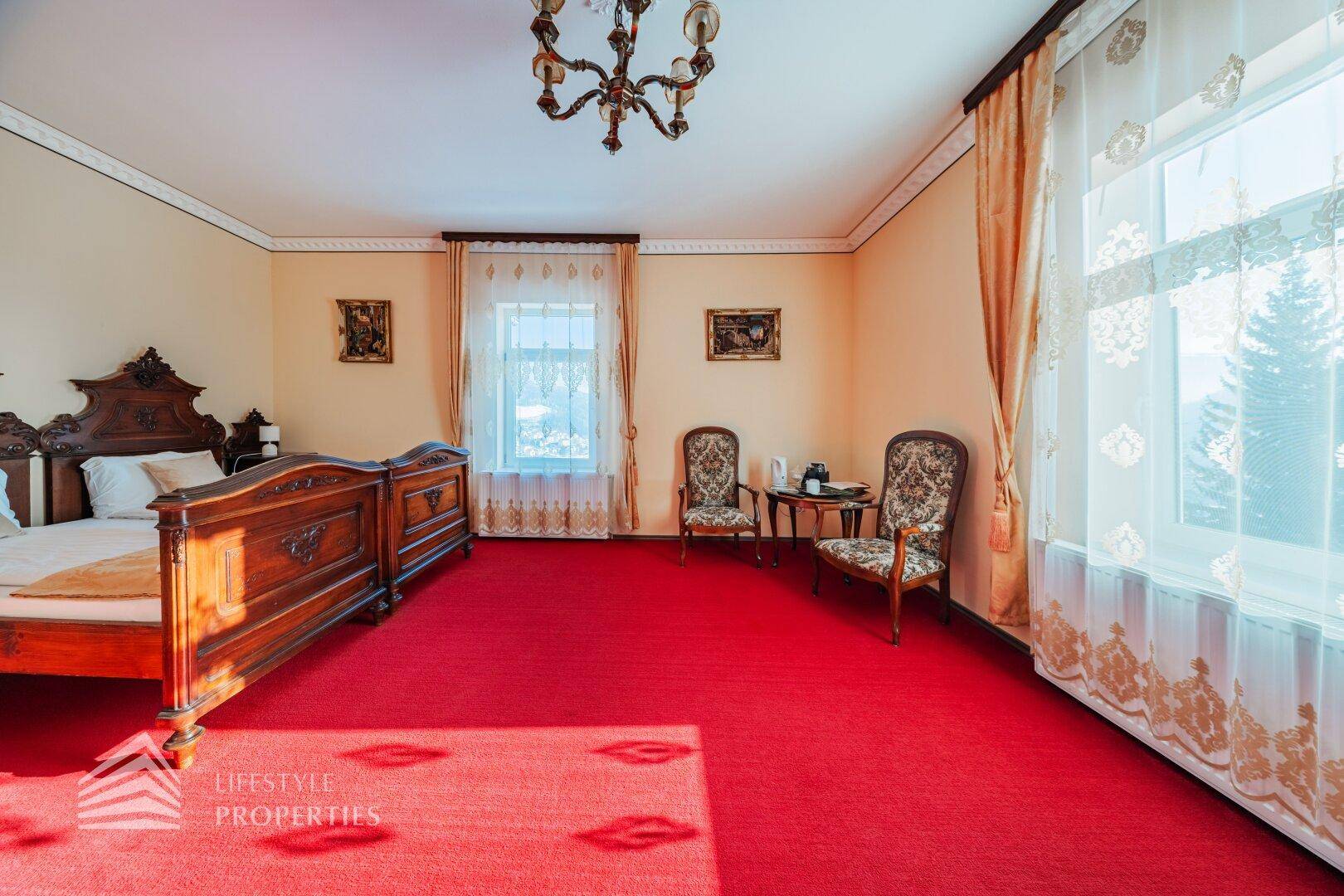 Helles Schlafzimmer mit rotem Teppich, traditionellen Möbeln und Fenstern mit Blick ins Grüne.