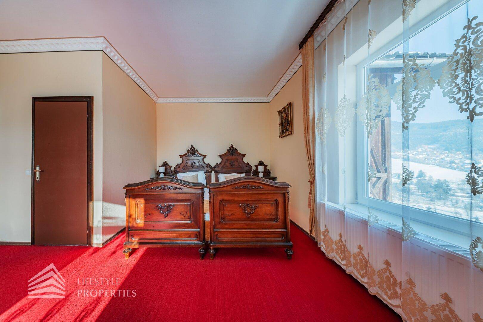 Schlafzimmer mit rotem Teppich, zwei Einzelbetten aus dunklem Holz und Fenster mit Weitblick.