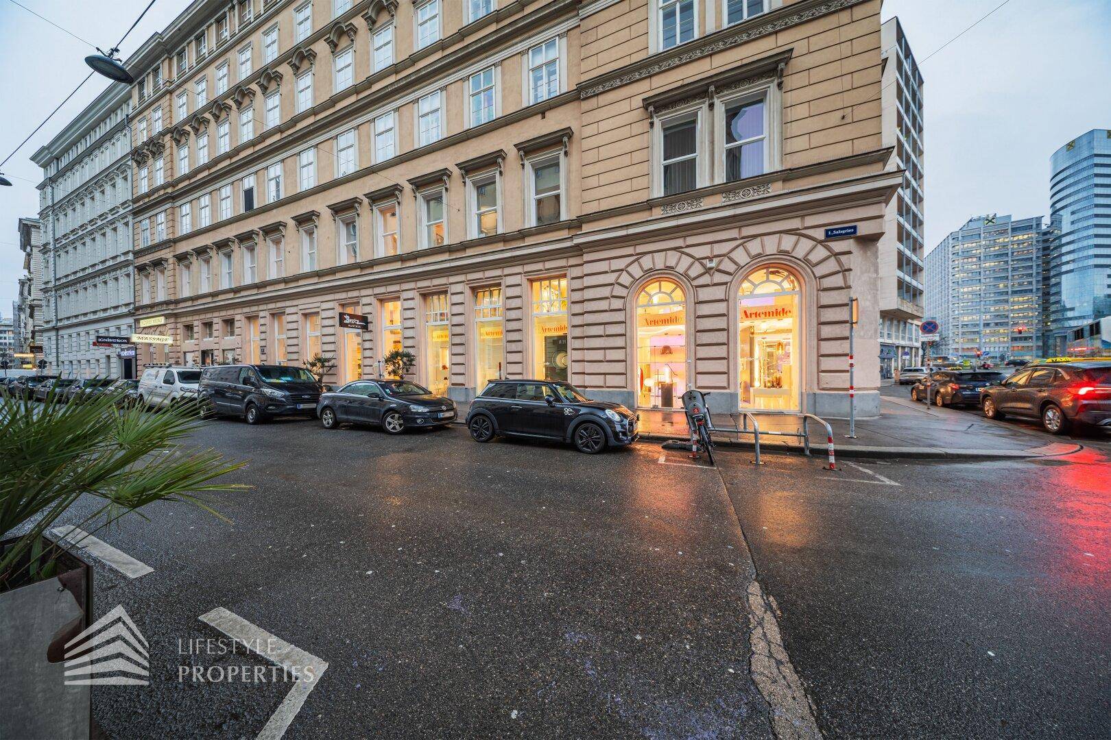 Historisches Gebäude mit modernen Ladenfronten an einer belebten Stadtstraße mit parkenden Autos.