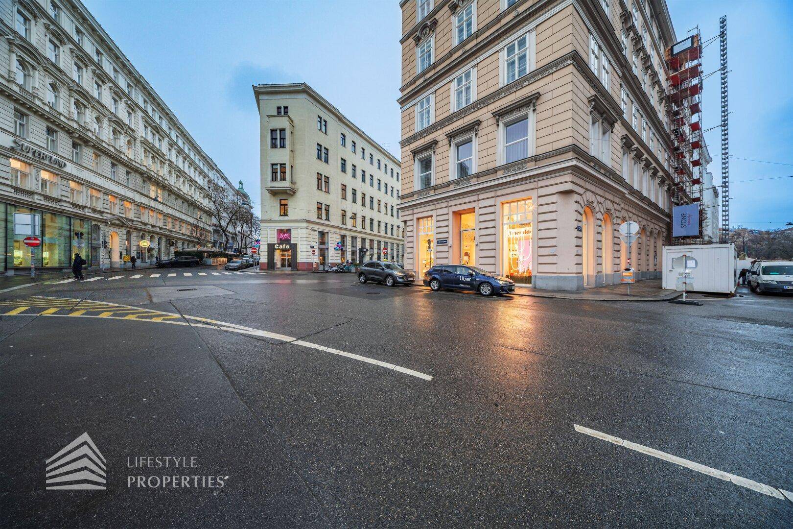 Stadtansicht mit historischen Gebäuden, belebter Kreuzung und parkenden Autos unter einem bewölkten Himmel.