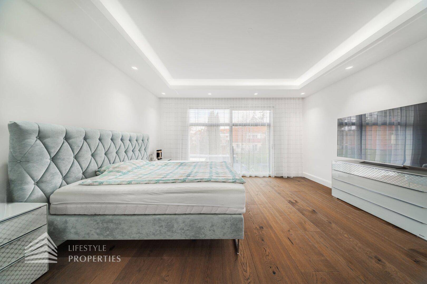 Geräumiges Schlafzimmer mit großem Bett, Holzboden und viel Tageslicht durch breite Fenster.